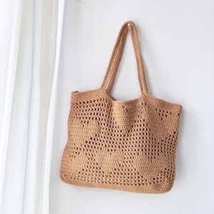 Op de afbeelding: Een bruin gehaakte tote bag met lange handvatten hangt aan een witte muur. De tas heeft een open geweven ontwerp. De tas is leeg en lijkt gemaakt van een natuurlijke vezel.
