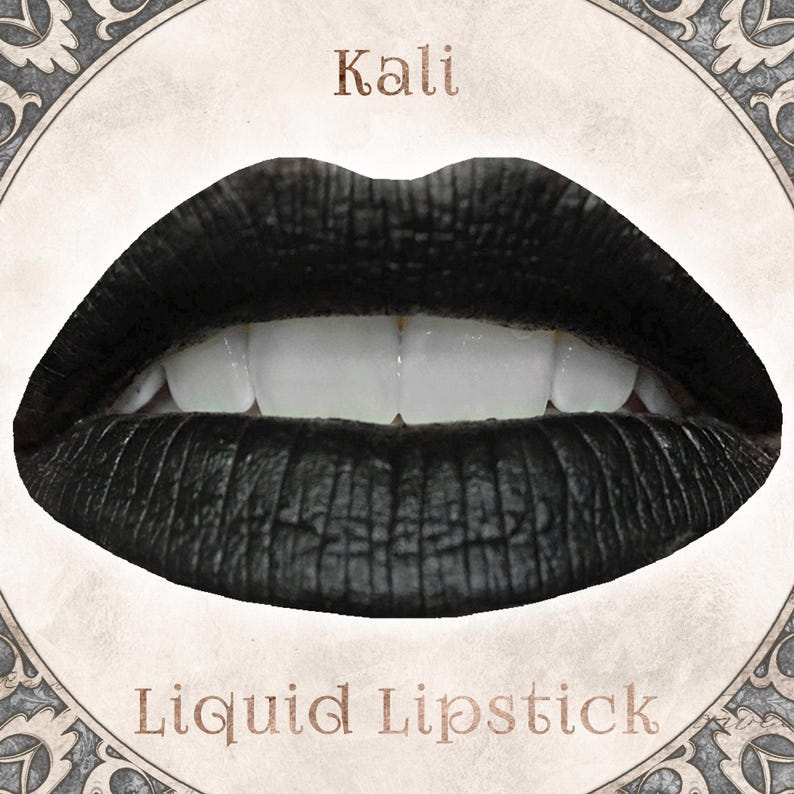 May include: A pair of lips coated in matte black liquid lipstick, showing a textured finish and white teeth. The word Kali is printed above the lips, and the words Liquid Lipstick are printed below, all set within a circular decorative frame.
