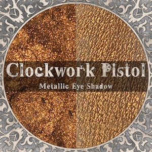 May include: A circular container of Clockwork Pistol metallic eye shadow featuring a split-screen display. The left side shows a deep bronze glitter texture, while the right side displays a smooth, metallic gold pigment with a fine, skin-like finish.