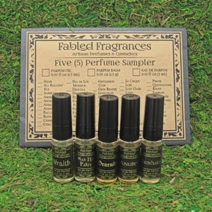 May include: Five small glass bottles with black labels and silver caps. The labels feature the words "Fabled Fragrances" and the names of the perfumes: "Wraith", "Mad Tea Party", "Dracula", "Anubis", and "Samhain". The bottles are arranged in a row on a green mossy surface. The bottles are part of a perfume sampler set.