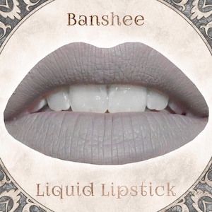 May include: A pair of lips coated in a matte, light grey liquid lipstick. The word Banshee is printed in brown above the lips, and the words Liquid Lipstick are printed in brown below the lips, all set within a circular, ornate frame.