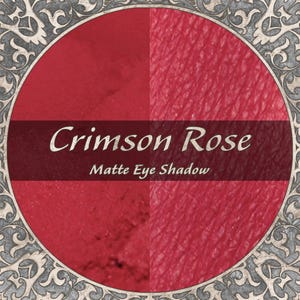 May include: A circular Crimson Rose matte eye shadow product display featuring a split-tone red background. The text Crimson Rose Matte Eye Shadow is printed in white across the centre. The circular frame has a decorative grey filigree border.