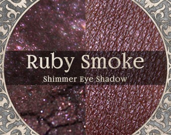 May include: A circular graphic featuring the text Ruby Smoke Shimmer Eye Shadow. The image displays a deep burgundy, glittery powder texture on the left and a smooth, metallic, dark reddish-brown swatch on the right, framed by an ornate border.