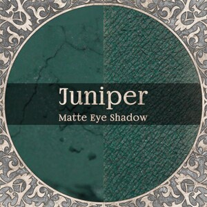 以下が含まれることがあります： Juniperとラベル付けされたダークグリーンのマットアイシャドウを特徴とする円形のグラフィック。デザインには、中央の製品見本とJuniper Matte Eye Shadowというテキストを囲む、複雑なスクロールワーク模様の装飾的なシルバー調の境界線が含まれています。