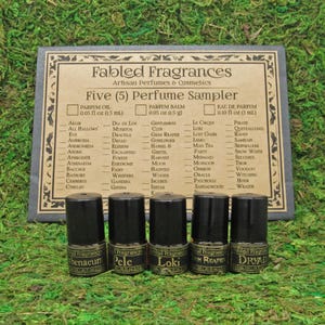 May include: Five small glass bottles with black caps and gold labels. The labels have the names "Aethernaeum", "Pele", "Loki", "Grim Reaper", and "Dryad" on them. The bottles are arranged in a row on a green mossy surface. The bottles are part of a perfume sampler set. The set is called "Fabled Fragrances Five (5) Perfume Sampler".