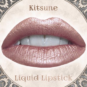 Op de afbeelding: Een paar lippen bedekt met een metallic ros&eacute;gouden vloeibare lippenstift. De tekst Kitsune staat boven de lippen en de tekst Liquid Lipstick staat eronder. De afbeelding is omlijst door een cirkelvormige rand met versierde hoekdetails.
