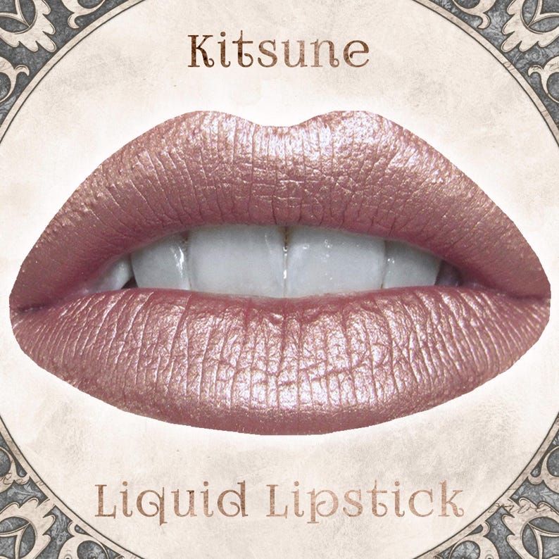Op de afbeelding: Een paar lippen bedekt met een metallic ros&eacute;gouden vloeibare lippenstift. De tekst Kitsune staat boven de lippen en de tekst Liquid Lipstick staat eronder. De afbeelding is omlijst door een cirkelvormige rand met versierde hoekdetails.