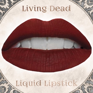 May include: A pair of lips coated in a deep, matte dark red liquid lipstick, showing white teeth. The image features the text Living Dead above the lips and Liquid Lipstick below, all framed within a circular border with ornate gray corner details.