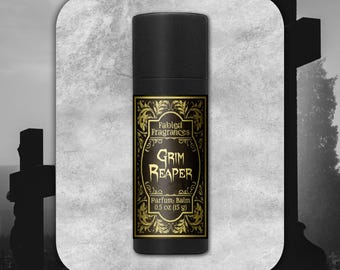 May include: A black cylindrical tube of Fabled Fragrances Grim Reaper parfum balm. The label features gold ornate filigree borders, black text, and states 0.5 oz (15 g). The container is centered against a light gray textured background.