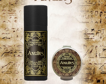 May include: A black cylindrical tube and a small round tin of Fabled Fragrances Anubis parfum balm. The tube label reads 0.5 oz (15 g) and the tin label reads 0.05 oz (1.5 g). Both containers feature gold decorative borders and black text.