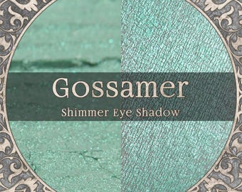 May include: A circular frame with an ornate gray border surrounds a split-view of mint green shimmer eye shadow. The text Gossamer Shimmer Eye Shadow is centered across the middle of the image in a dark gray horizontal band.