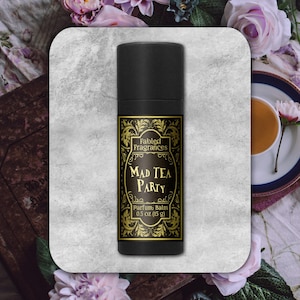 May include: A black cylindrical tube of Fabled Fragrances Mad Tea Party parfum balm. The label features gold ornate filigree borders on a black background with the text Fabled Fragrances, Mad Tea Party, and the weight of 0.5 oz (15 g).