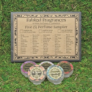 以下が含まれることがあります： Fabled Fragrancesの香水サンプルセット。5種類の香りが含まれています。「Succubus」や「All Hallows Eve」などのラベルが付いた、小さくて丸い容器が入っています。サンプルは緑色の苔の上に置かれています。