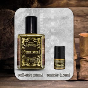 May include: A clear glass bottle of Fabled Fragrances Gunslinger parfum oil with a black cap, labeled 0.5 fl oz (15 mL), next to a smaller sample bottle labeled 0.05 fl oz (1.5 mL), both displayed against a ruler showing metric and imperial scales.