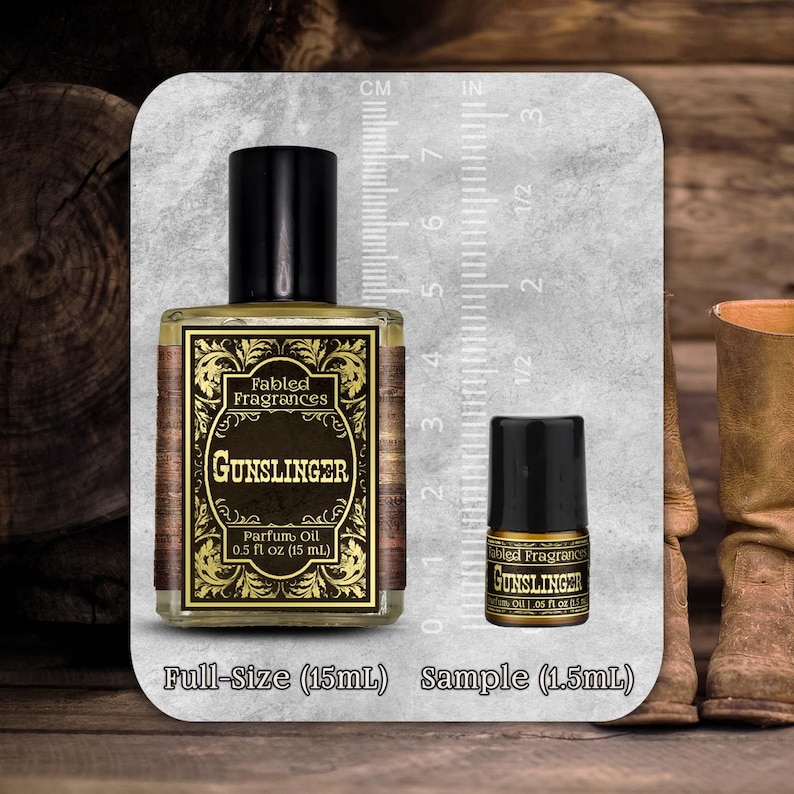 May include: A clear glass bottle of Fabled Fragrances Gunslinger parfum oil with a black cap, labeled 0.5 fl oz (15 mL), next to a smaller sample bottle labeled 0.05 fl oz (1.5 mL), both displayed against a ruler showing metric and imperial scales.