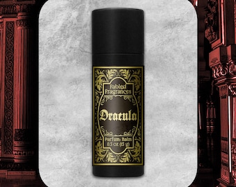 May include: A black cylindrical tube of Fabled Fragrances Dracula parfum balm. The label features gold ornate filigree borders on a black background with the text Fabled Fragrances, Dracula, and Parfum Balm 0.5 oz (15 g) printed in gold.