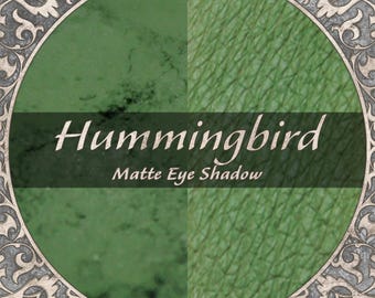May include: A circular graphic featuring a split-tone green background with a textured finish. The text Hummingbird Matte Eye Shadow is centered horizontally across the middle. The entire design is enclosed in an ornate, metallic-toned border.