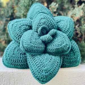 May include: A dark teal crochet cushion shaped like a succulent plant. The cushion features multiple layered, rounded, and pointed petals constructed with a dense yarn stitch pattern, creating a three-dimensional, textured, and sculptural appearance.