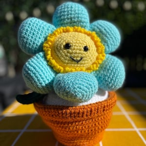 May include: A crocheted flower toy with light blue petals and a yellow centre featuring a stitched smiling face with black eyes. The flower is planted in a textured orange crocheted pot. The entire object is crafted from yarn with visible stitches.