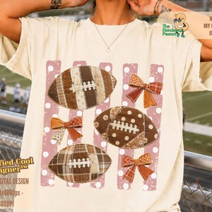Pode incluir: Uma t-shirt de manga curta cor de creme com uma impressão gráfica de três bolas de futebol americano castanhas com padrões de patchwork, três laços estampados laranja e castanhos e riscas verticais cor-de-rosa com bolas brancas.