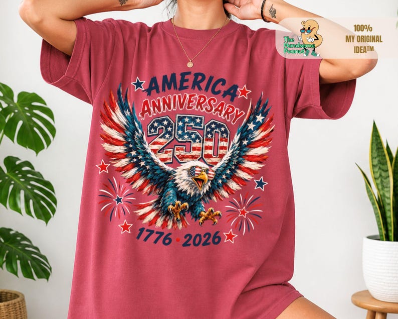 May include: A red short-sleeve t-shirt featuring a graphic of a bald eagle wearing sunglasses with a stars and stripes pattern. The text reads AMERICA ANNIVERSARY 250, 1776-2026, with red, white, and blue fireworks and star accents surrounding the eagle.