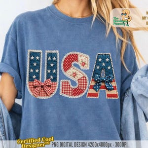 Op de afbeelding: Blauw T-shirt met het woord "USA" op de voorkant. De letters zijn versierd met patriottische patronen, waaronder sterren, strepen en strikken. Het shirt heeft opgerolde mouwen en een casual pasvorm.