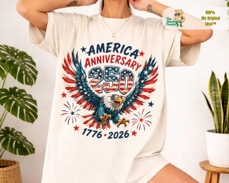 May include: A cream-colored t-shirt featuring a graphic of a bald eagle with red, white, and blue wings wearing sunglasses. The text reads AMERICA ANNIVERSARY 250, 1776-2026, surrounded by stars and fireworks. The design is centered on the chest.