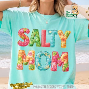 May include: A light turquoise t-shirt with the words "SALTY MOM" in colorful, floral-patterned letters. The shirt has short sleeves and is worn on a beach. The text "The Handsome Peanut" and "Certified Cool Designer" are also visible.