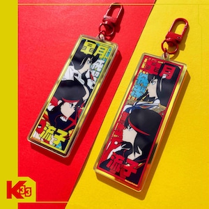 Puede incluir: Dos llaveros rectangulares con cierres rojos. Cada llavero presenta ilustraciones de estilo anime de personajes con cabello negro y detalles rojos. Texto japonés visible en ambos llaveros, sobre un fondo rojo, amarillo y azul.