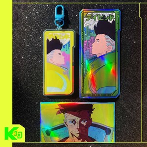 May include: Three holographic Cyberpunk 2077 themed items. A yellow keychain with a character profile, a vertical card with Japanese text and a character profile, and a rectangular sticker with a character portrait.