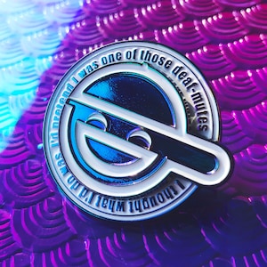 May include: A silver and blue enamel pin with the text "I thought what I'd do no was pretend I was one of those deaf-mutes." The pin features a stylized design with a smiling face and a horizontal bar.
