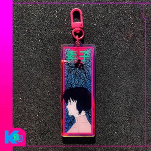 以下が含まれることがあります： ピンクの留め金が付いた長方形のアクリルキーホルダー。キーホルダーには、黒髪の人の横顔と日本語のテキストが描かれています。背景は青と黒のデザインです。