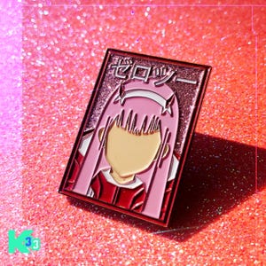 May include: A rectangular pink enamel pin featuring a stylized anime character with pink hair and a red outfit. The pin has Japanese text at the top and is set against a glittery pink background.