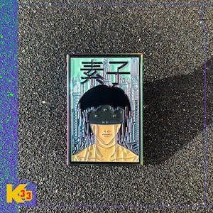 May include: A rectangular enamel pin with a rainbow metallic border featuring a stylized illustration of a character with dark hair and a visor over their eyes. The background depicts a city skyline in light blue, with black Japanese text at the top.