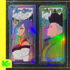 May include: Two holographic stickers with anime-style characters. One shows a woman with blue hair, the other a man in a green jacket. Both have Japanese text and cityscapes. The stickers have a rainbow sheen and are rectangular.