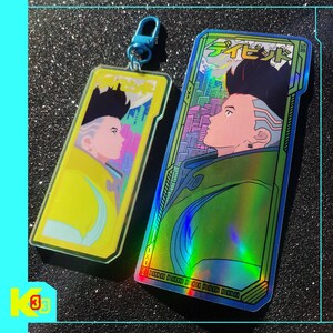 May include: Two rectangular keychains and stickers with a futuristic design. One is yellow, the other holographic, both featuring a side profile of a character with black hair and Japanese text.