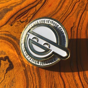 May include: A round, gold-colored pin with a white and black design. The pin features a stylized smiling face and the text "I thought what I'd do was pretend I was one of those deaf-mutes."