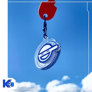 May include: A circular clear acrylic keychain featuring a blue graphic of a smiling face with a cap. The keychain is attached to a dark blue lobster clasp and a red key head. The design includes white text printed along the circular edge.