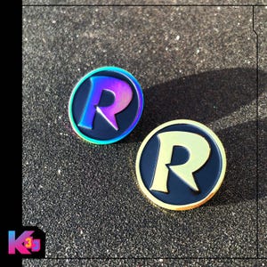 May include: Two circular enamel pins featuring a stylised letter R in the centre. One pin has a black background with a gold-toned letter R and gold border. The other pin has a black background with an iridescent, rainbow-coloured letter R and border.