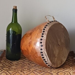 May include: A green glass bottle with a cork stopper stands next to a large, brown gourd drum. The drum has a light-colored rim with black tacks and a small handle. The objects rest on a patterned, brown surface.
