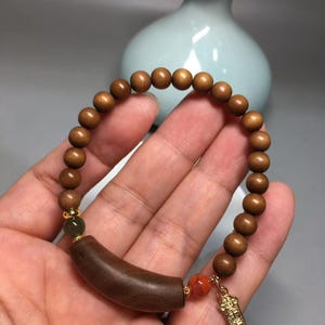 Puede incluir: Una pulsera de cuentas con esferas de madera marrón lisa, un cilindro de madera marrón oscuro curvado, una cuenta pequeña de color verde oscuro, una cuenta naranja moteada y un dije de metal dorado con texto grabado, unidos por espaciadores dorados.