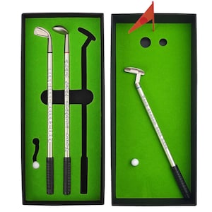 Puede incluir: Un set de bolígrafos de golf en dos cajas negras con inserciones de fieltro verde. El set incluye tres bolígrafos plateados con forma de palo de golf y empuñaduras negras, con el texto Your custom text here, una bola blanca y una bandera roja.