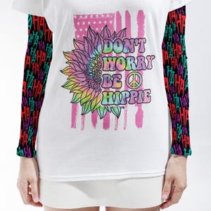 May include: A white t-shirt with long sleeves featuring a black background with repeating colorful "HA" text. The front graphic shows a tie-dye sunflower, a pink American flag design, and the text "DON'T WORRY BE HIPPIE" with a peace sign.