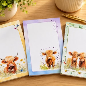 May include: Three rectangular notepads featuring illustrations of Highland cattle. The left pad has a yellow border with a single cow, the center pad has a purple border with a single cow, and the right pad has a green floral border with two cows.