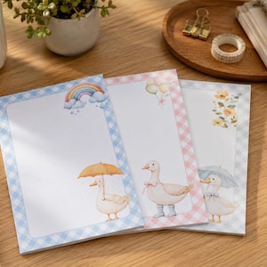 May include: Three rectangular stationery sheets featuring gingham borders in blue, pink, and light blue. The designs include watercolor illustrations of ducks with umbrellas, a rainbow with clouds, balloons, and yellow floral accents on white paper.