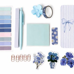 May include: A stationery set featuring a light blue pen, a lavender highlighter, a stack of light blue sticky notes, a blue flower hair clip, patterned washi tape, a striped bookmark with a bow, floral stickers, and copper heart paper clips.