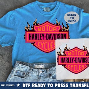 May include: A bright blue t-shirt featuring a graphic of a Harley-Davidson logo in pink and orange, accented with a leopard print flame design. The text reads MOTOR HARLEY-DAVIDSON CYCLES. A separate transfer sheet shows the same graphic design.