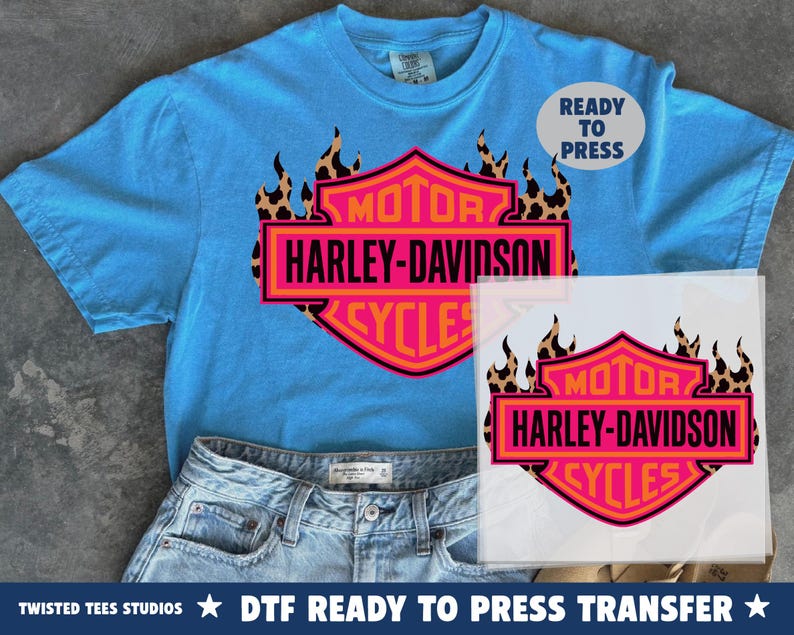 May include: A bright blue t-shirt featuring a graphic of a Harley-Davidson logo in pink and orange, accented with a leopard print flame design. The text reads MOTOR HARLEY-DAVIDSON CYCLES. A separate transfer sheet shows the same graphic design.