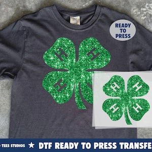 May include: Dark gray t-shirt with a large, sparkling green four-leaf clover design. The clover has the letter "H" in each leaf. A "Ready to Press" sticker is on the shirt. A DTF ready to press transfer is also visible.