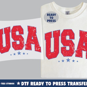 May include: A white t-shirt with the word "USA" in distressed red lettering and blue outlines. Below the text are three blue stars. A "Ready to Press" sticker is on the shirt. A matching transfer is on the left.